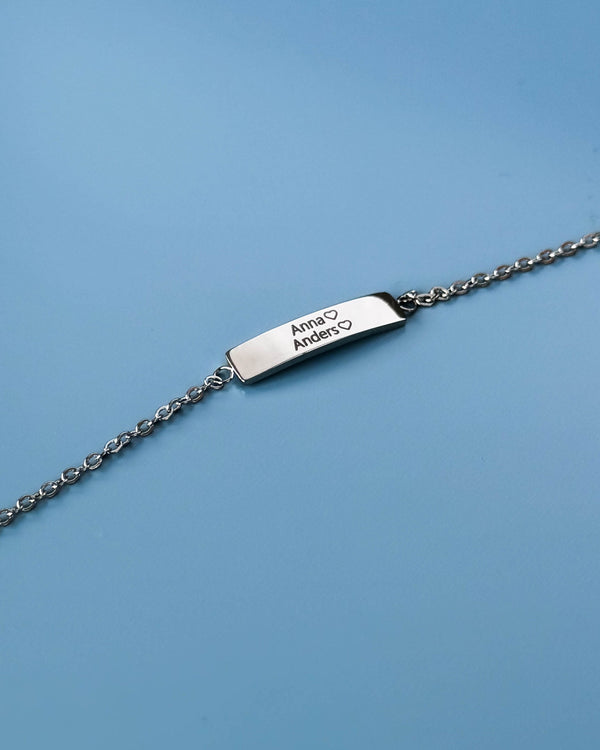 EVI name bracelet in silver-coloured stainless steel with the names "Anna" ♡ "Anders" ♡ engraved, against a light blue background