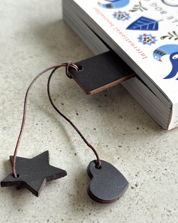 PAGE bookmark in dark brown leather tucked into a book, with leather heart and star charms hanging out on a cord