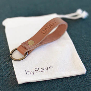 White fabric gift bag with byRavn logo and cognac leather keyring with the engraving "Daddy" on a green surface