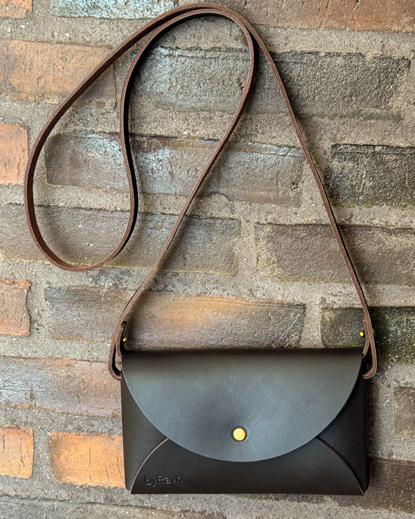 INA shoulder bag in dark brown leather with long strap hanging on a red brick wall