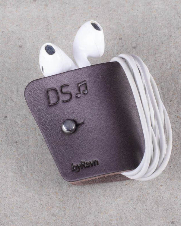 KAX earbud clip in dark brown leather with white earpods wrapped around and the engraving "DS" with a music note, on concrete