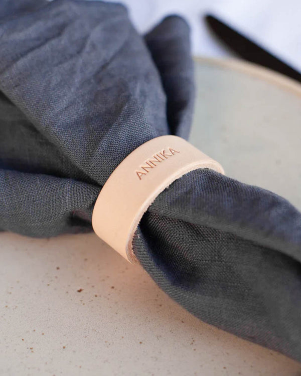 KNOT napkin ring in natural leather engraved with the name "Annika" around a navy blue cloth napkin on a plate