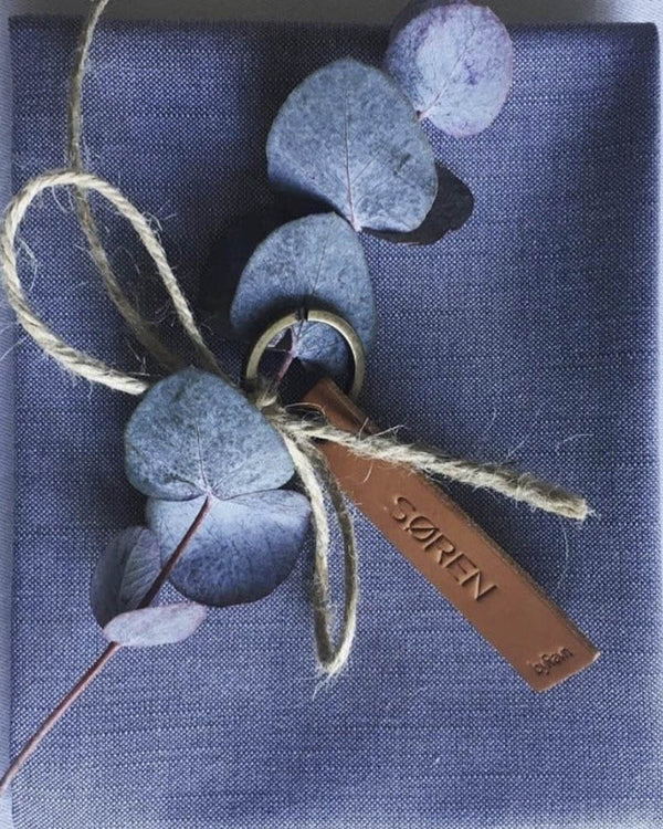 MERRY leather place card in cognac with the name "Søren" engraved, tied with twine and eucalyptus on a blue napkin