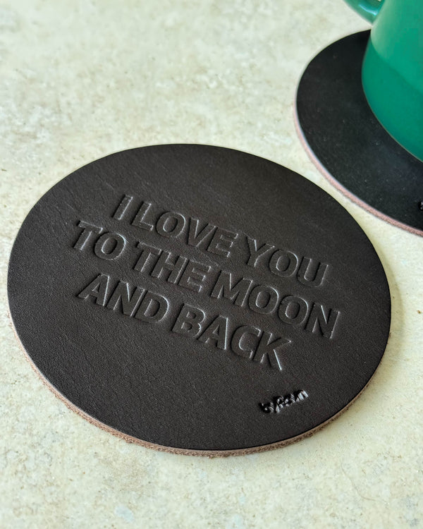 MOKKA coaster in dark brown leather embossed "I love you to the moon and back" on a light table next to a green cup