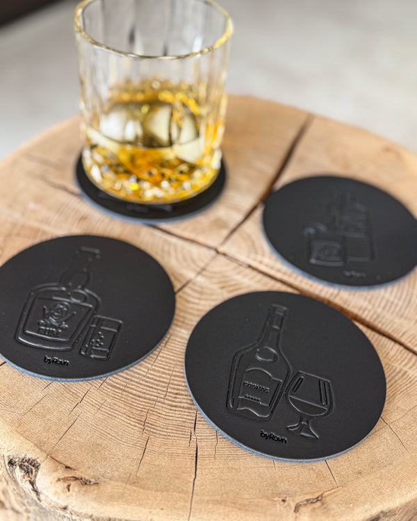 MOKKA spirits coasters in black leather embossed with rum, cognac and gin on a wooden slab by a crystal glass of whisky