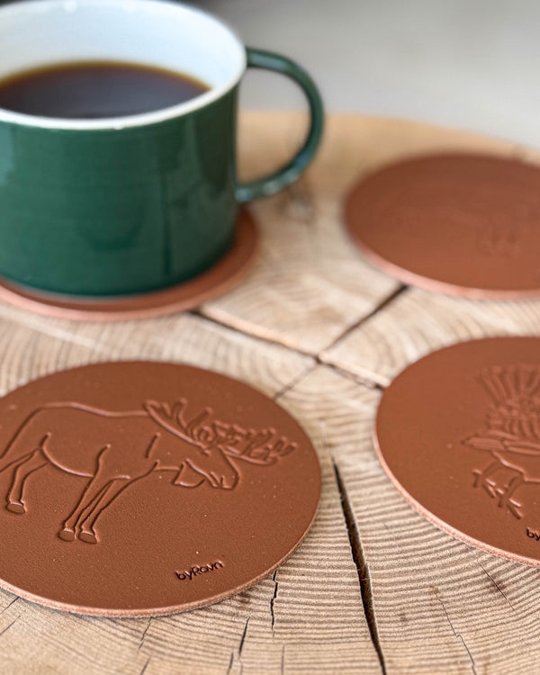 MOKKA hunting coasters in cognac leather embossed with elk and capercaillie on a wooden slab next to a green coffee cup on a coaster