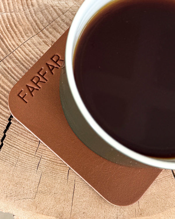 MOKKA square coaster in cognac leather with the engraving "Farfar" under a cup of coffee on a wooden slice