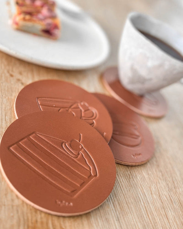 MOKKA coffee & cake coasters in cognac leather embossed with layer cake, next to coffee cup and plate of cake on a wooden table