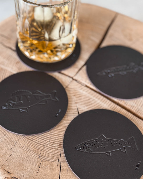 MOKKA angler coasters in dark brown leather embossed with sea trout, perch and pike on a wooden slab by a whisky glass