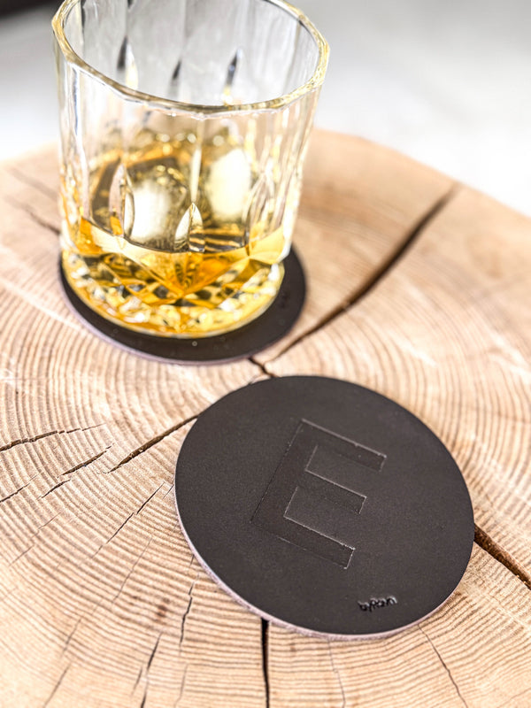 MOKKA coaster in dark brown leather embossed with the letter "E" on a wooden slab next to a whisky glass on a coaster