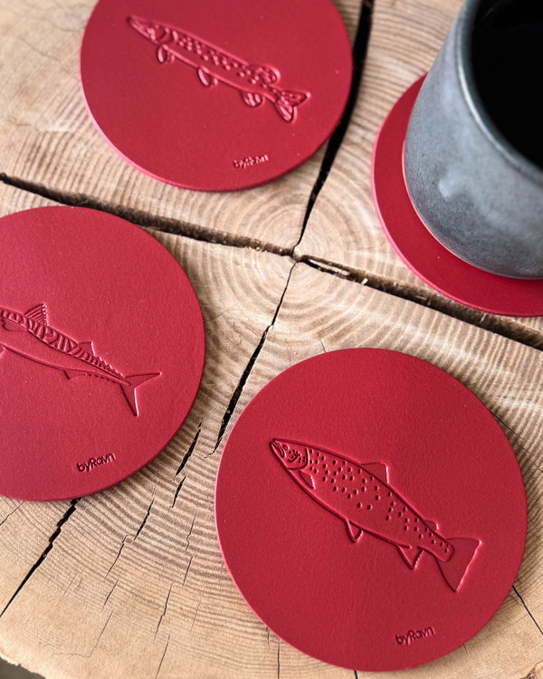 MOKKA angler coasters in red leather embossed with sea trout, mackerel and pike on a wooden slab next to a grey coffee cup