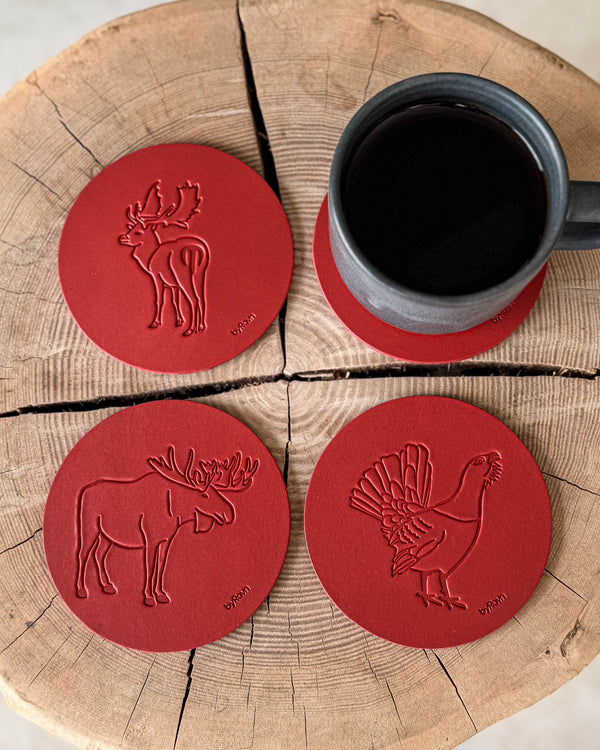 MOKKA hunting coasters in red leather embossed with elk, capercaillie and fallow deer on a wooden slab by a grey coffee cup on a coaster