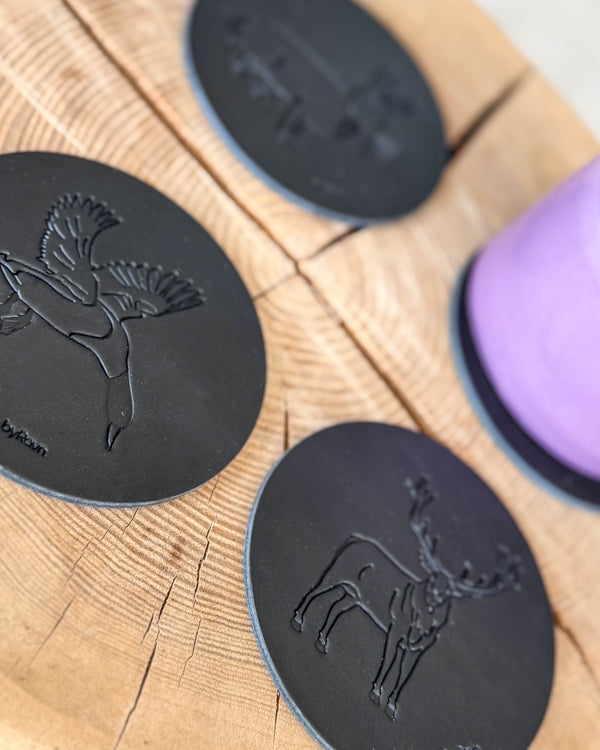 MOKKA hunting coasters in black leather embossed with flying mallard and red deer stag on a wooden slab next to a purple cup