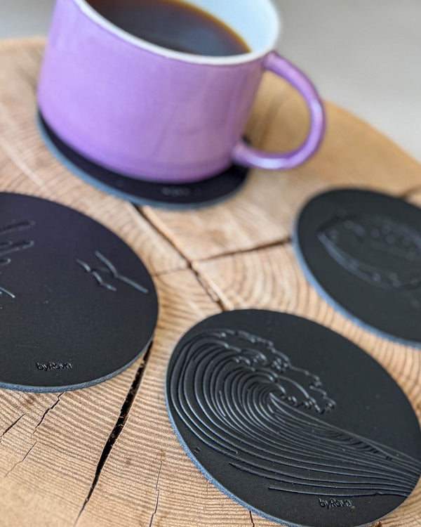 MOKKA summer house coasters in black leather embossed with wave, marram grass and flatfish on a wooden slab with purple coffee cup