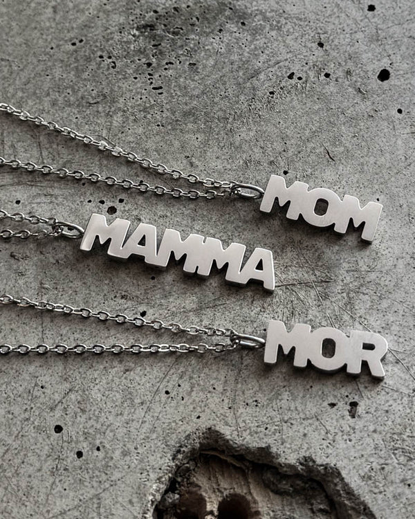 MOM necklaces in silver-coloured stainless steel with letter pendants "MOM", "MAMMA" and "MOR", resting on raw concrete
