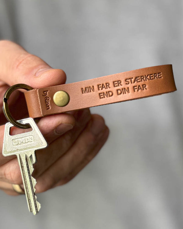 RAVN leather keyring in cognac with the engraving "Min far er stærkere end din far", held in hand with key