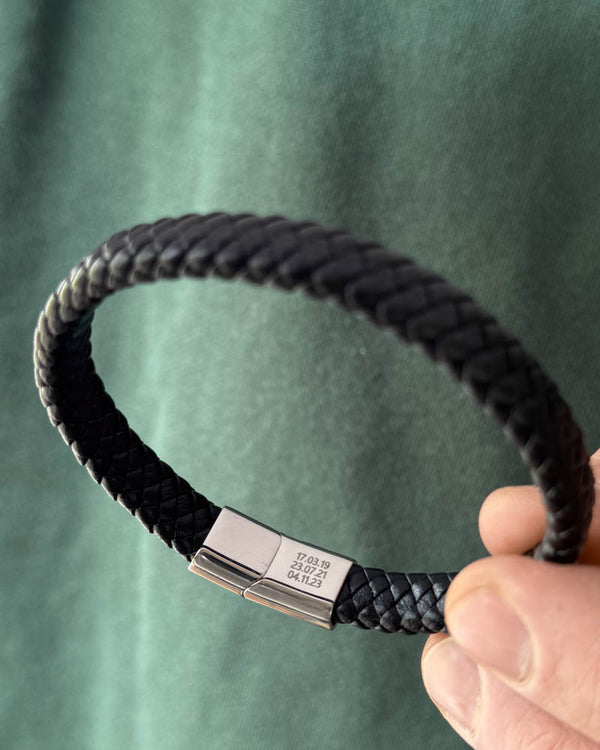 RAY leather bracelet in black braided leather held in hand, the dates "17.03.19, 23.07.21, 04.11.23" engraved on the clasp
