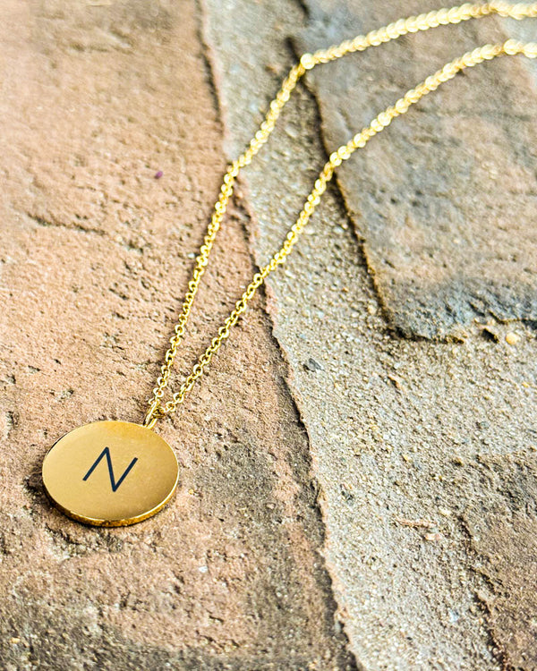 SIA LETTER necklace in gold-plated stainless steel with the letter "N" engraved on a round pendant, lying on reddish tiles
