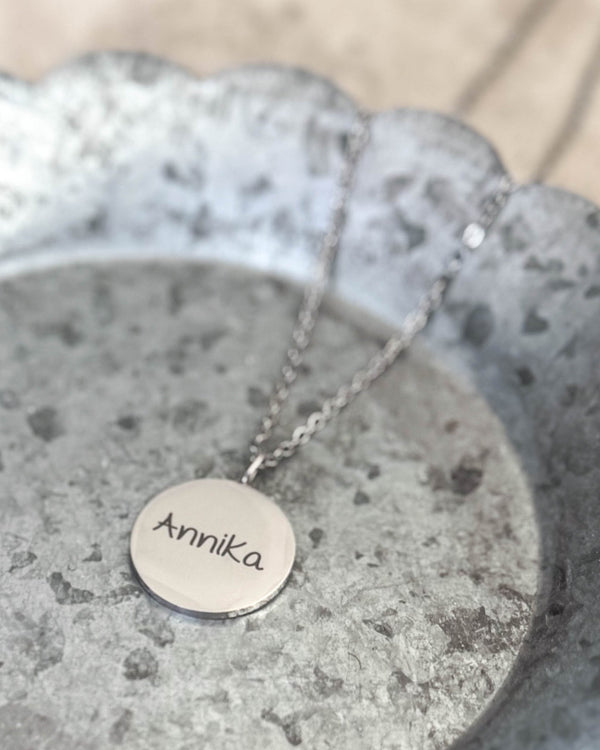 SIA NOTE SCRIPT name necklace in silver-coloured stainless steel with the name "Annika" engraved in handwriting, lying on a zinc tray