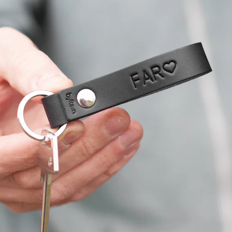 Julegave til far – sort RAVN lædernøglering med præget "Far" ♡ og sølvfarvet nøglering, holdt i hånden
