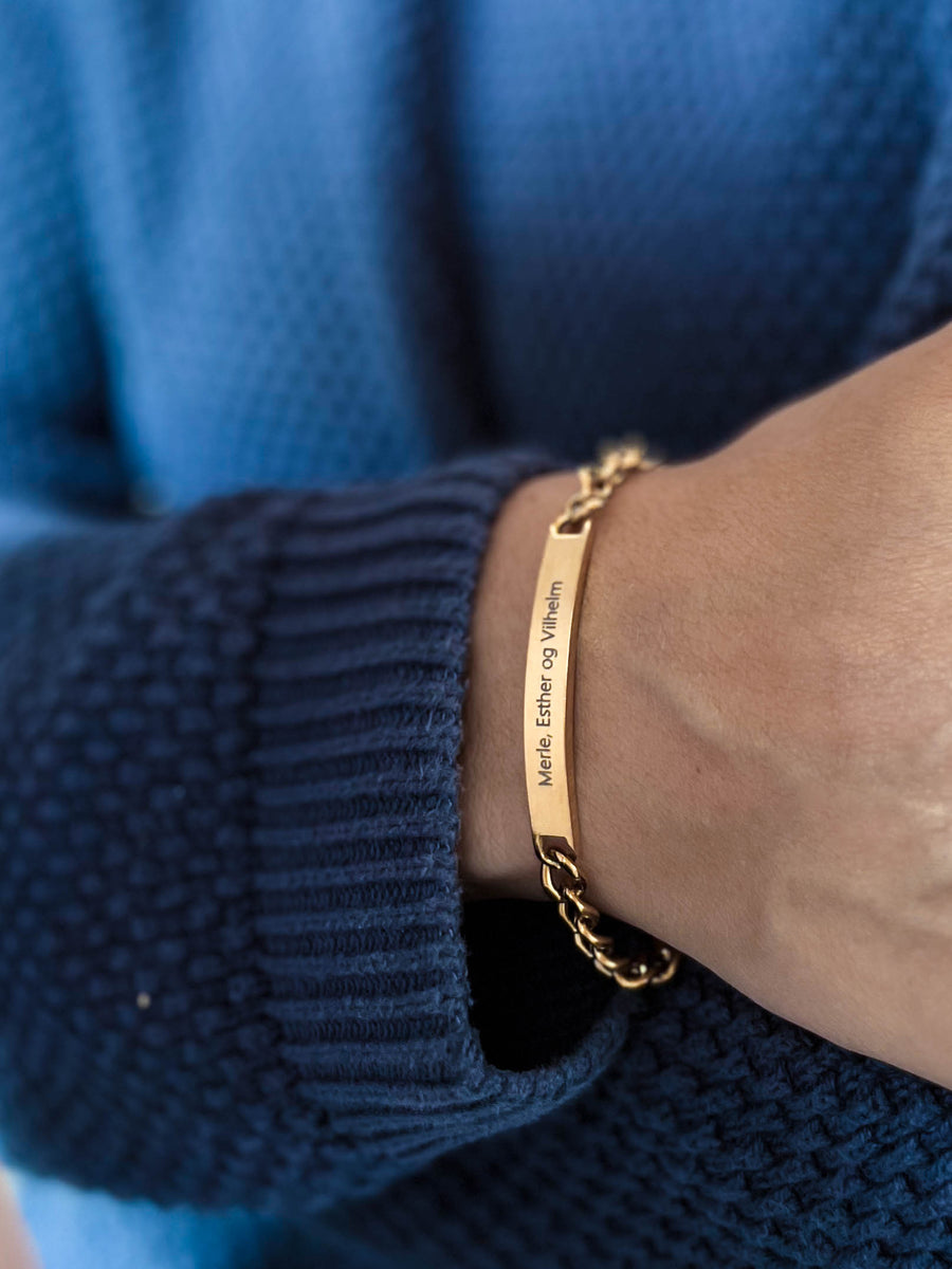AVA name bracelet in gold-plated stainless steel with "Merle, Esther og Vilhelm" engraved, worn on the wrist with a blue knit