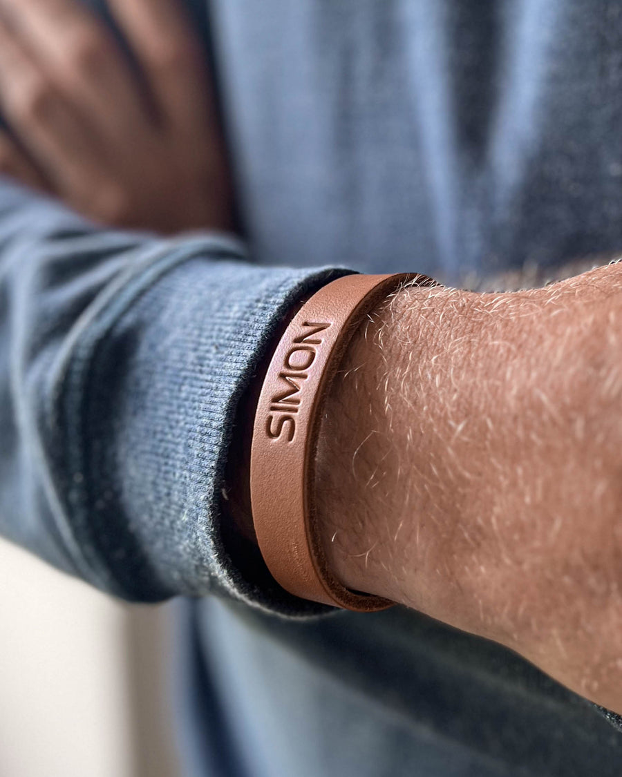 MONO leather bracelet in cognac with the name "Simon" engraved, worn on the wrist of a man in a blue knit