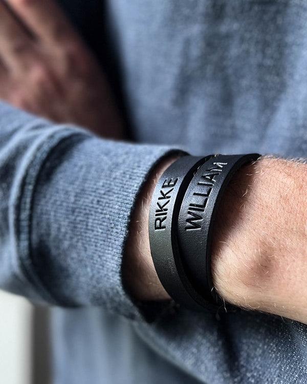 DUO leather bracelet in black with the names "Rikke" and "William" engraved, worn on a man's wrist in a grey knit
