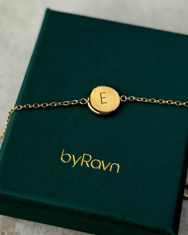 IBI LETTER bracelet in gold-plated stainless steel with the letter "E" engraved, lying on a dark green byRavn jewellery box