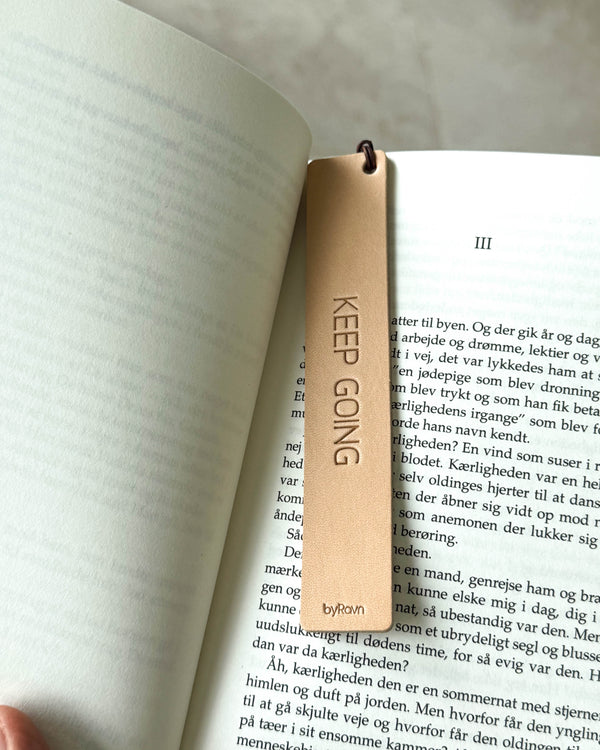PAGE bookmark in natural leather with the engraving "Keep going" placed between the pages of an open book