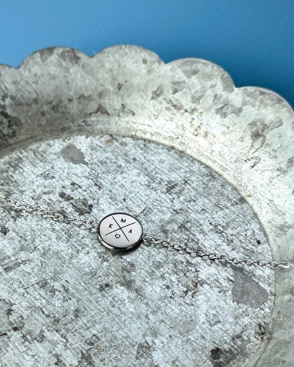 IBI X bracelet in silver-coloured stainless steel with cross, initials "K", "M", "A" and a heart engraved, on a zinc tray, blue background