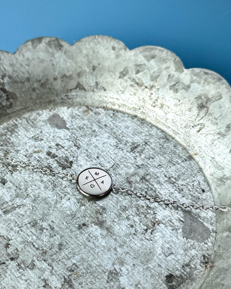 IBI X bracelet in silver-coloured stainless steel with cross, initials "K", "M", "A" and a heart engraved, on a zinc tray, blue background