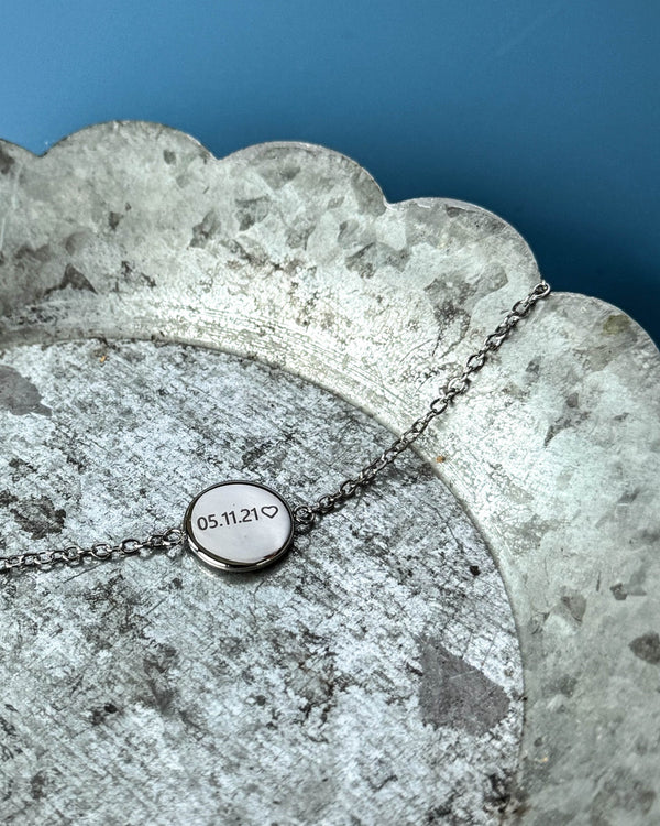 IBI NOTE bracelet in silver-coloured stainless steel with "05.11.21" ♡ engraved on a round disc, on a zinc tray against a blue background