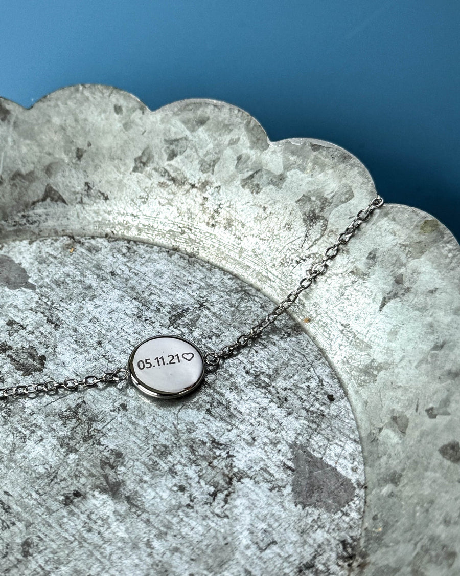 IBI NOTE bracelet in silver-coloured stainless steel with "05.11.21" ♡ engraved on a round disc, on a zinc tray against a blue background