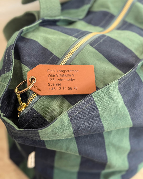 ATLAS leather luggage tag, here in cognac, with engraved address "Pippi Langstrømpe, Villa Villakulla 9" on a green-blue striped bag