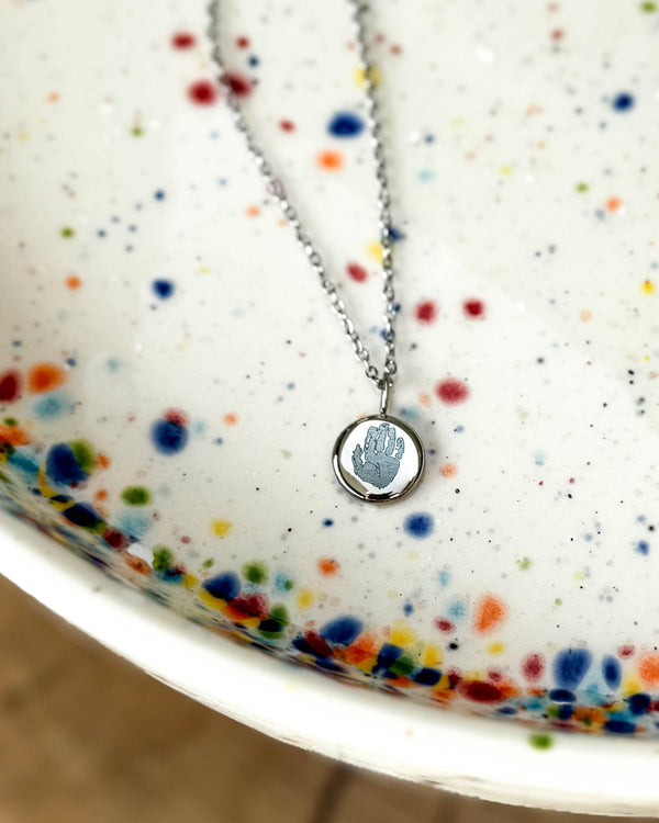 AYA MOTIF necklace in silver-coloured stainless steel with a small handprint engraved on a round pendant, on a speckled ceramic dish