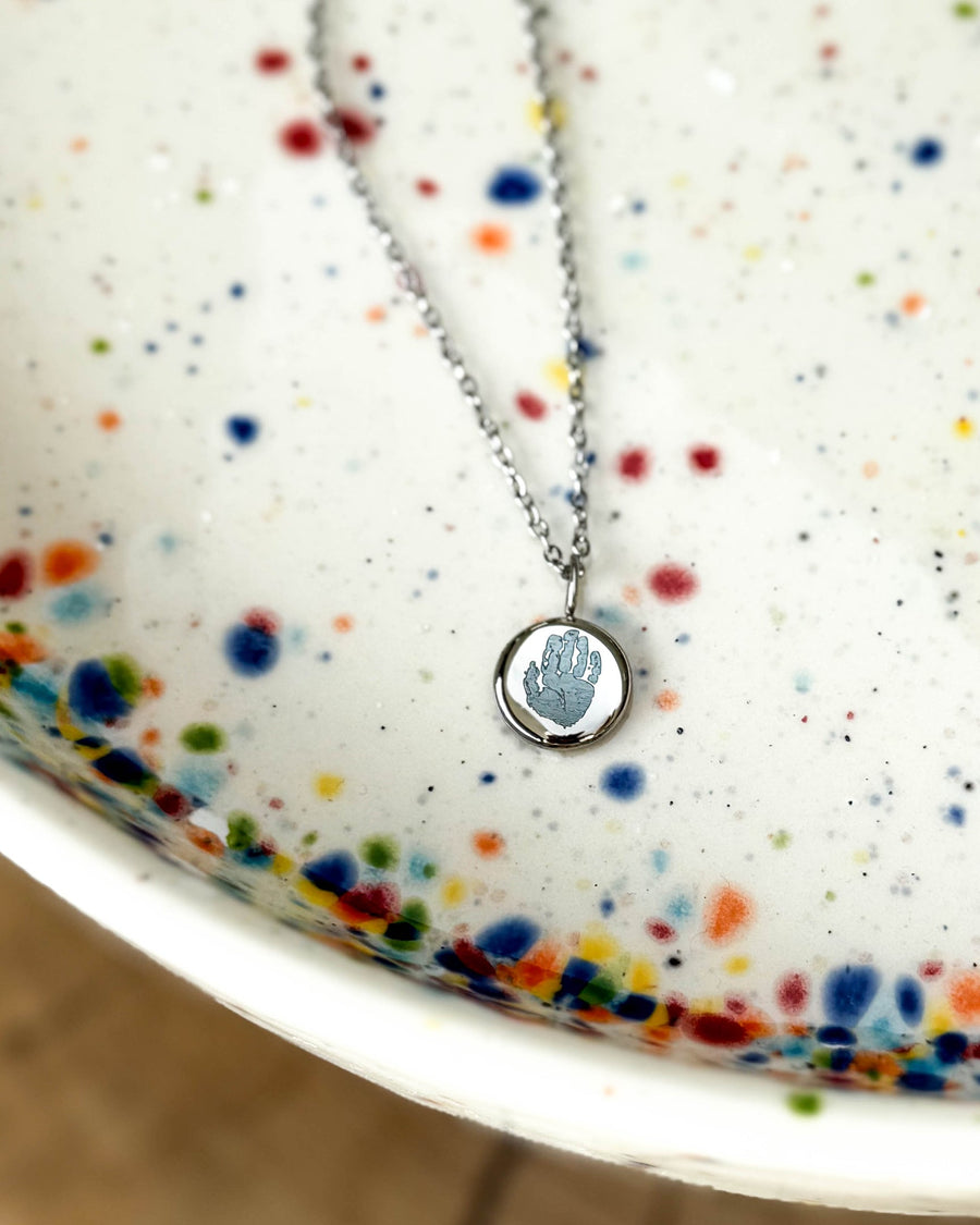 AYA MOTIF necklace in silver-coloured stainless steel with a small handprint engraved on a round pendant, on a speckled ceramic dish