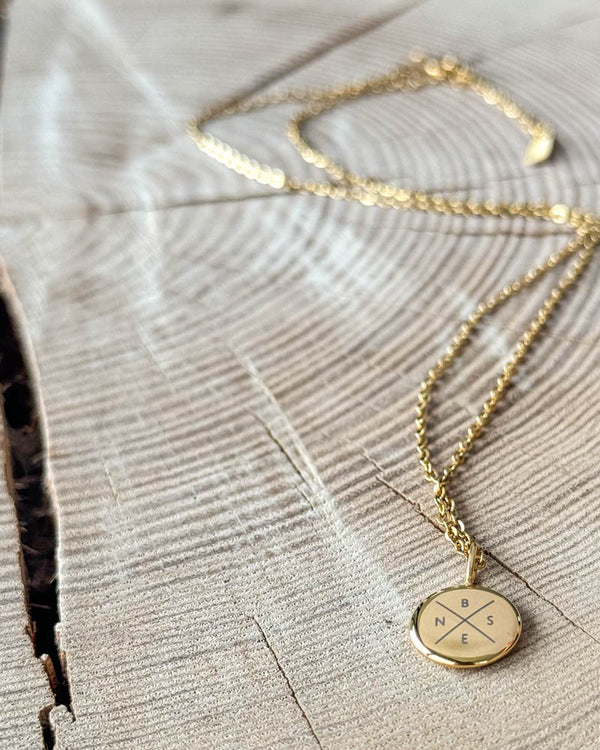 AYA X necklace in gold-plated steel with the initials "B N S E" engraved in a cross on a small round pendant, on light wood with a crack