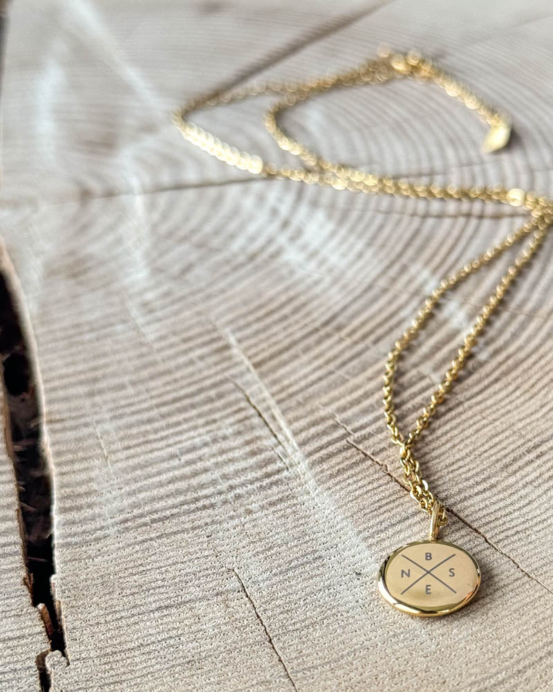 AYA X necklace in gold-plated steel with the initials "B N S E" engraved in a cross on a small round pendant, on light wood with a crack