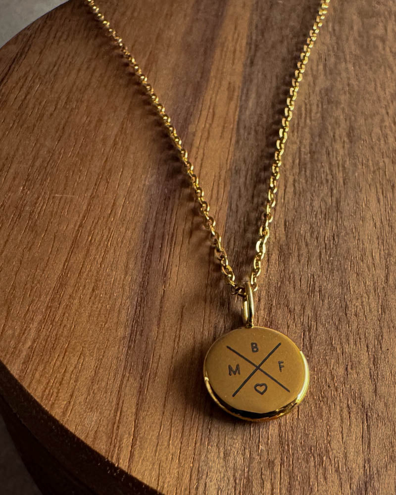AYA X necklace in gold-plated stainless steel with the letters "B M F ♡" engraved in a cross on a small pendant, on a dark wooden board