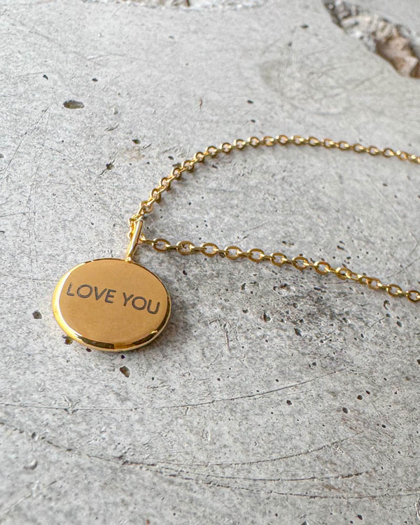 AYA NOTE necklace in gold-plated stainless steel with the engraving "Love you" on a small round pendant, lying on concrete