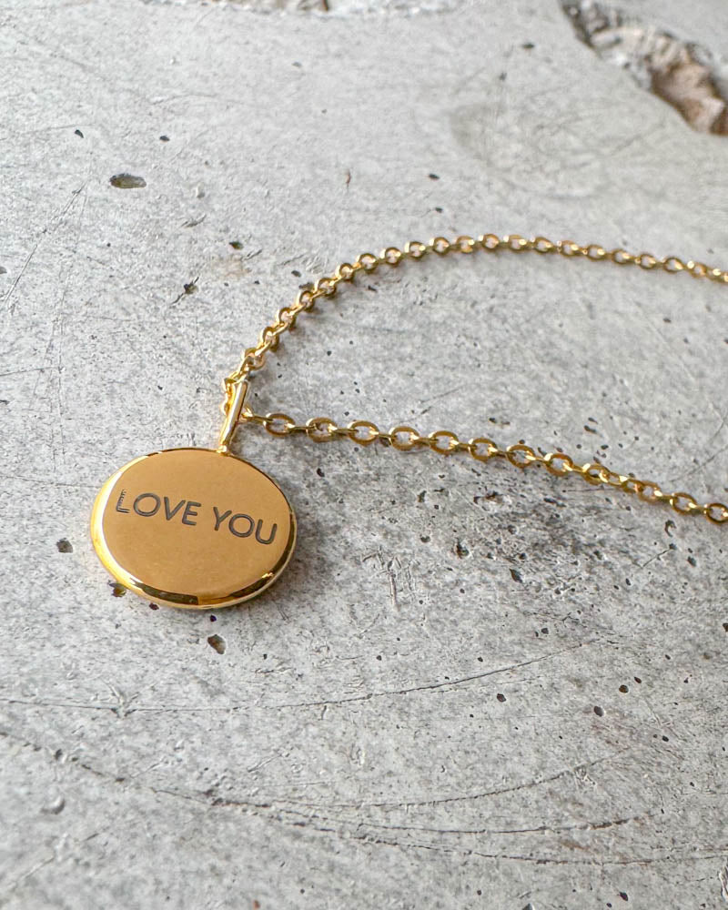 AYA NOTE necklace in gold-plated stainless steel with the engraving "Love you" on a small round pendant, lying on concrete