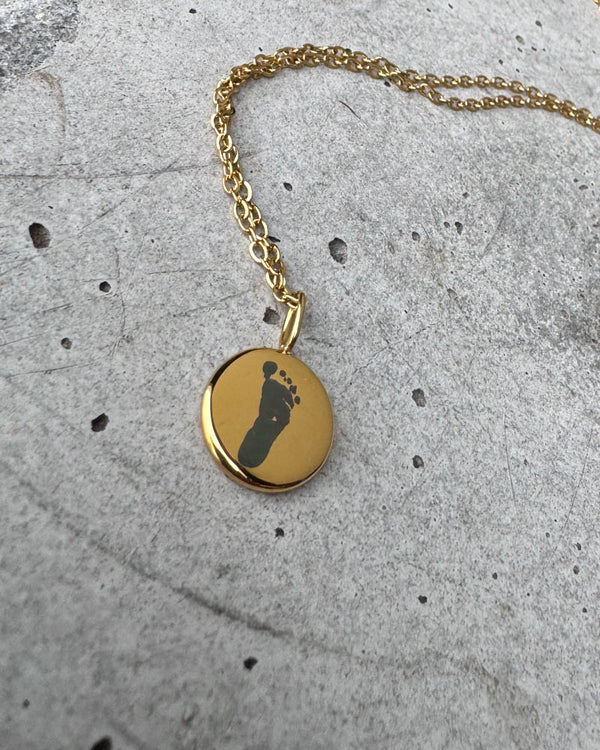 AYA MOTIF necklace in gold-plated stainless steel with a small footprint engraved on a round pendant, lying on concrete