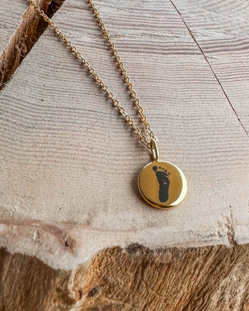 AYA MOTIF necklace in gold-plated stainless steel with baby's footprint engraved on a small pendant, on light wood with a crack