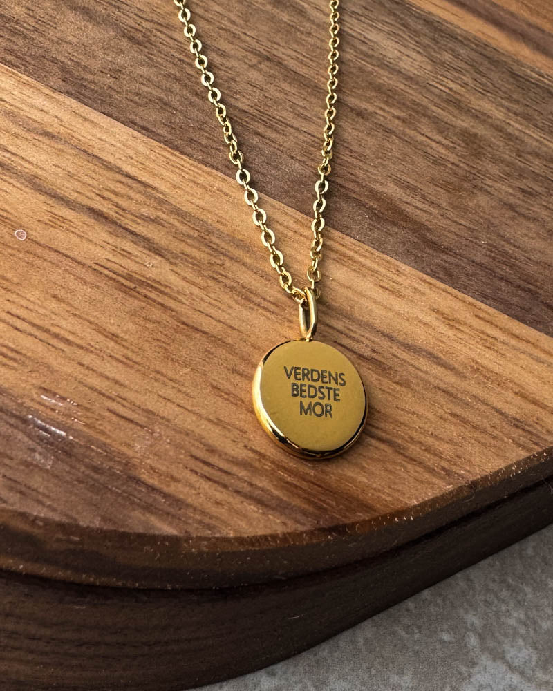 AYA QUOTE necklace with the engraving "Verdens bedste mor" on a small round pendant, shown here in gold-plated steel on a dark wooden board