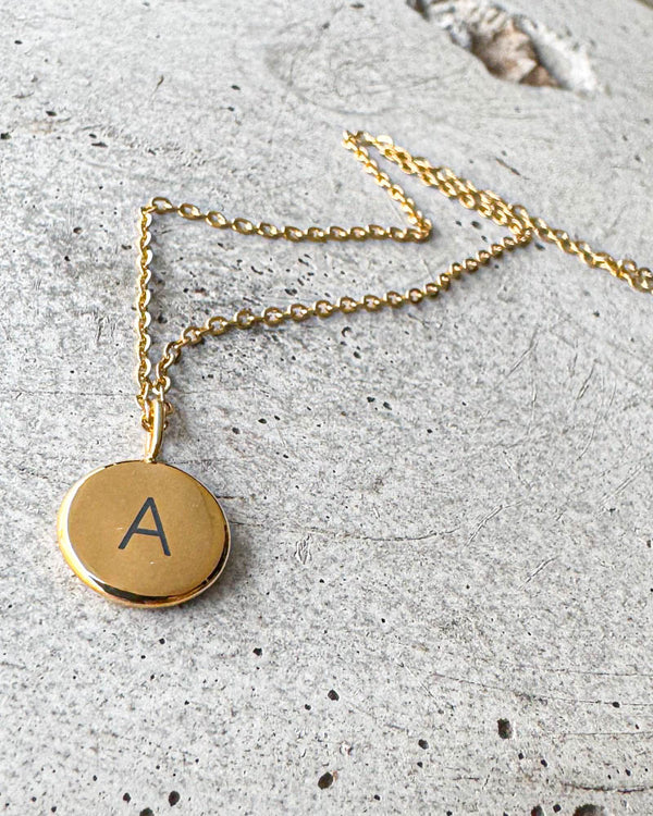 AYA LETTER necklace in gold-plated stainless steel with the letter "A" engraved on a small round pendant, lying on concrete