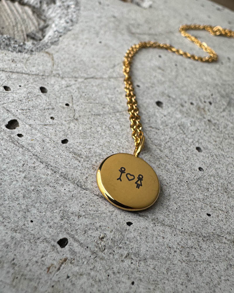 AYA STICK necklace in gold-plated steel with matchstick couple and heart engraved on a small round pendant, lying on concrete
