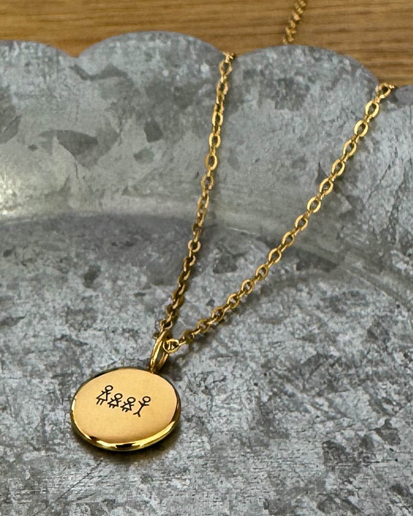 AYA STICK necklace in gold-plated steel with a matchstick family of four engraved on a small round pendant, lying on a zinc tray