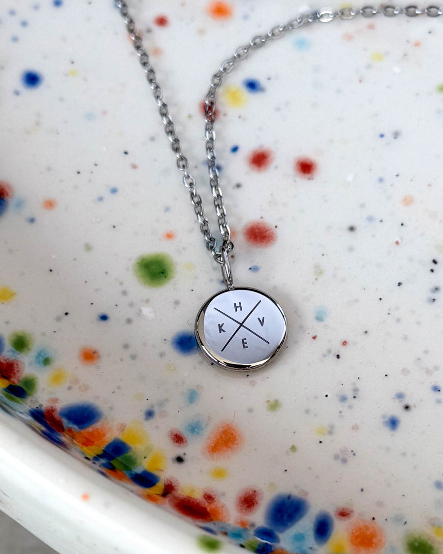 AYA X necklace in silver-coloured steel with the initials "H K V E" engraved in a cross on a small pendant, on a speckled ceramic dish
