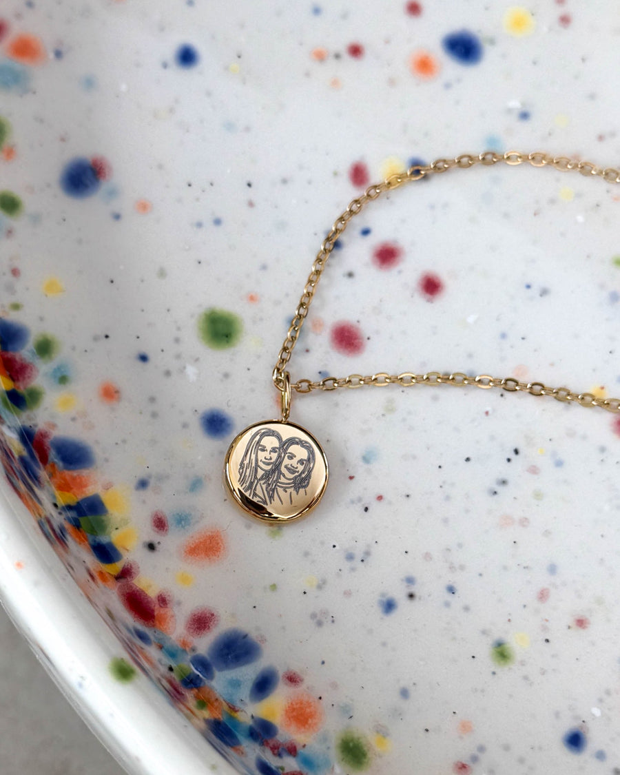 AYA PORTRAIT necklace in gold-plated steel with line-drawn portrait of two women on a small pendant, on a speckled ceramic dish