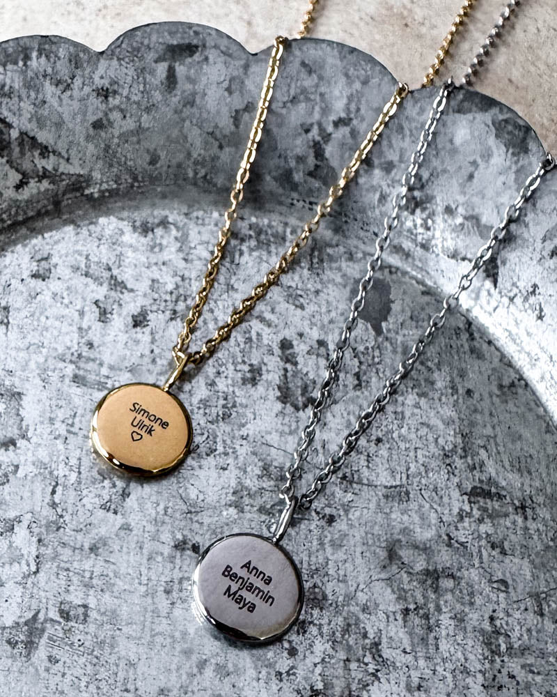 AYA QUOTE name necklaces in gold-plated and silver-coloured steel engraved with "Simone Ulrik" ♡ and "Anna Benjamin Maya", on a zinc tray